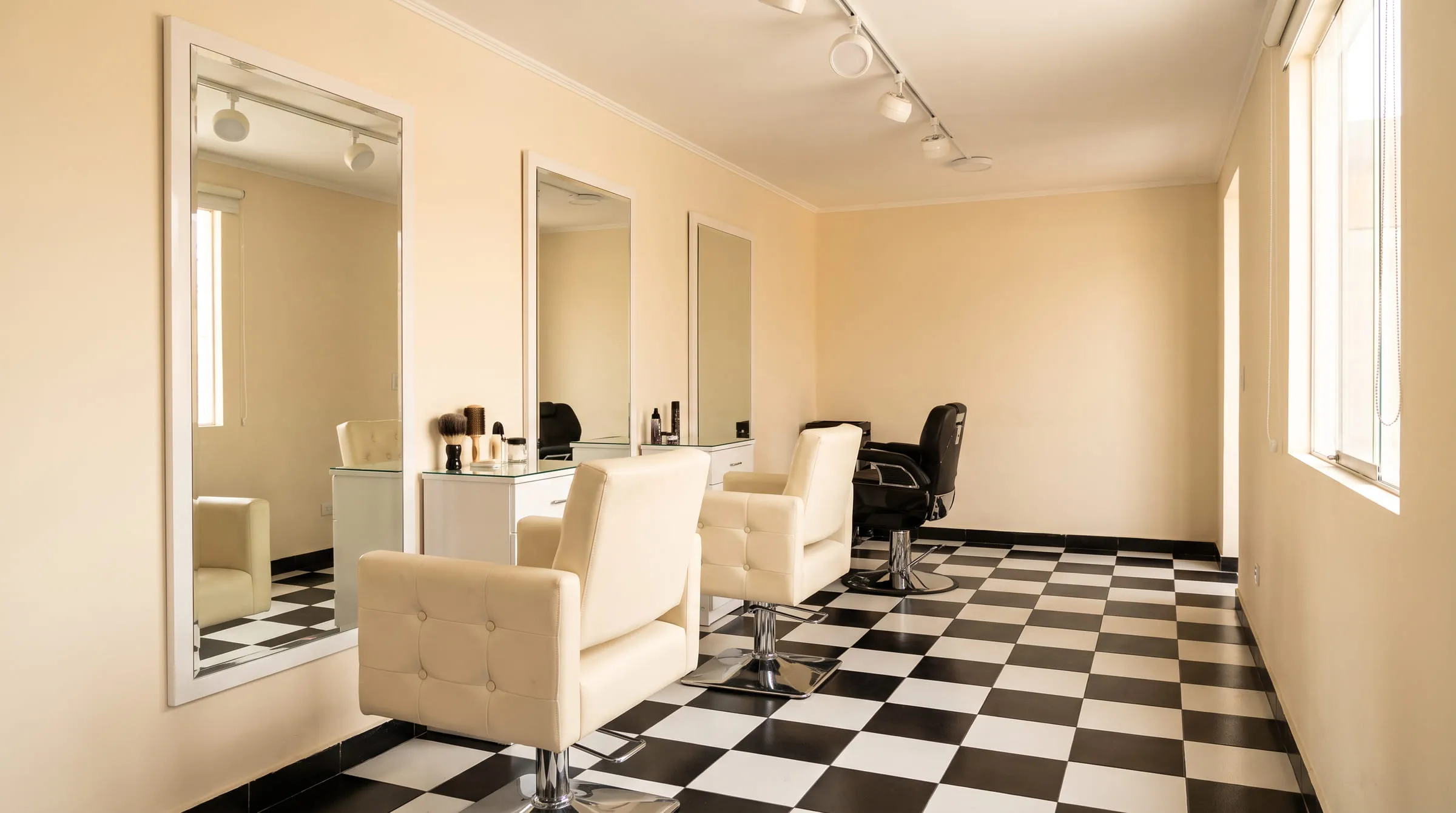 Interior del salón: fila de espejos de marco blanco, sillones capitoné en crema con base cromada y piso de damero negro y blanco, con luz natural cálida