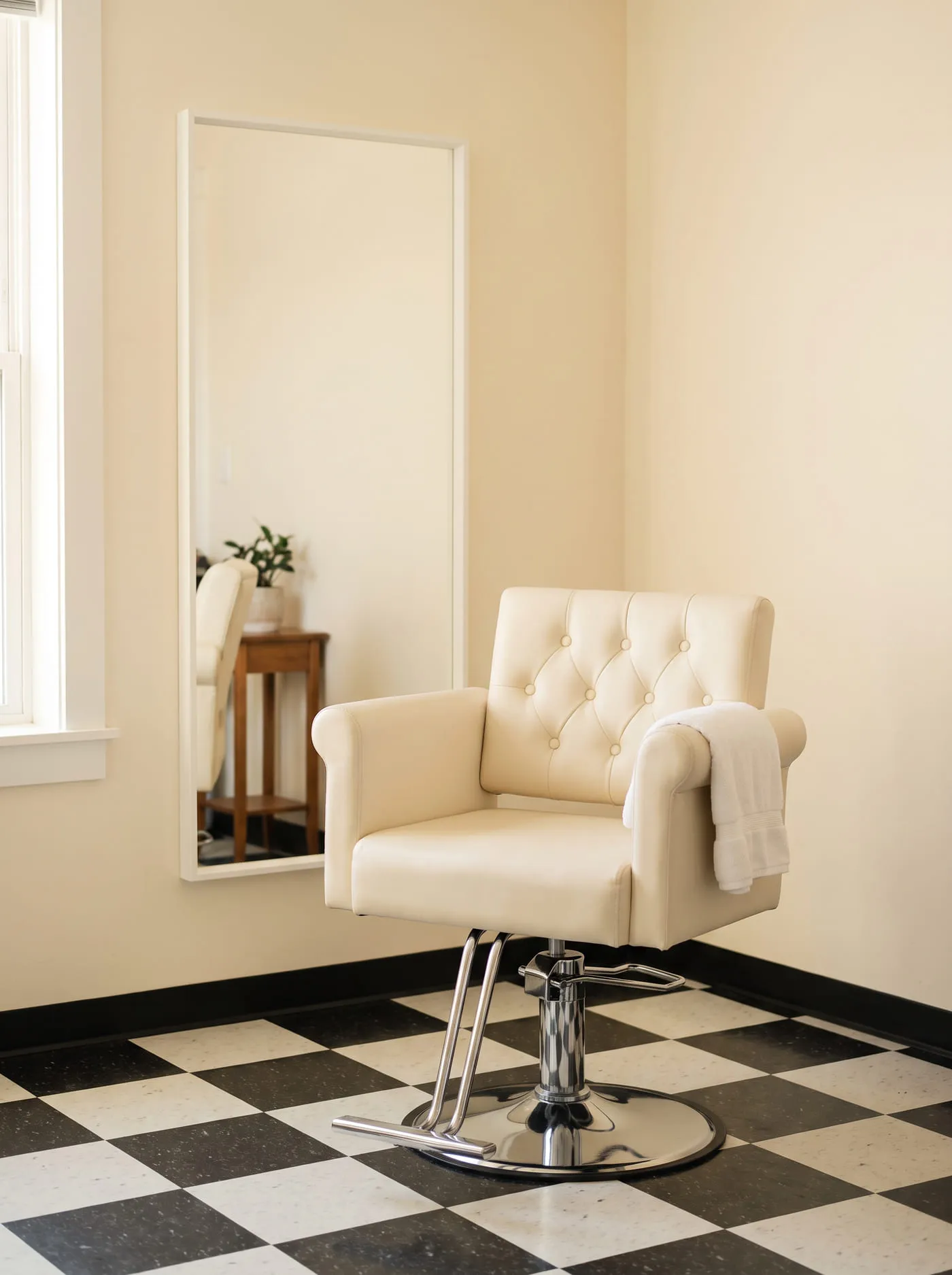 Un rincón del salón: sillón capitoné en crema con base cromada frente a un espejo de marco blanco, sobre el piso de damero negro y blanco, con luz natural cálida